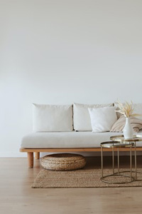 Dans un salon blanc, un canapé en bois aux larges coussins blancs est placé derrière des tables gigognes en métal doré. Sur le plateau, on voit un vase en porcelaine blanche